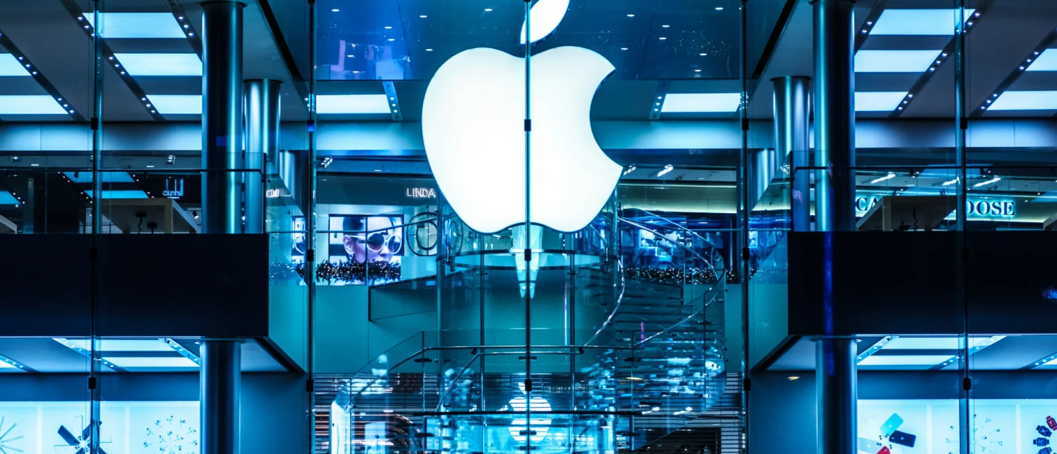 Apple-Logo an einer verglasten Fassade mit blau beleuchtetem Store im Hintergrund.