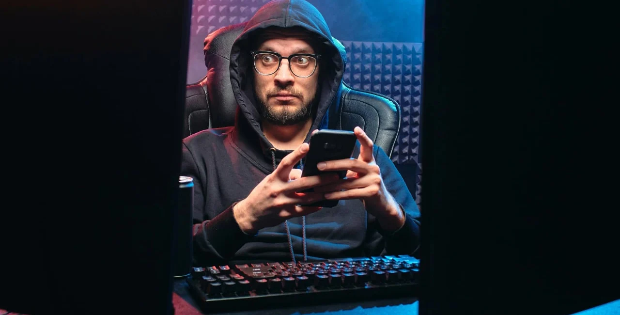 IT-Sicherheit und Cybersecurity: Ein Mann mit Brille und Kapuzenpullover sitzt vor zwei Monitoren in einem abgedunkelten Raum mit schalldämmenden Wänden. Er hält ein Smartphone und blickt überrascht auf den Bildschirm. Die Szene mit blauen und roten Lichtakzenten vermittelt eine Hacking- oder Cyberangriff-Atmosphäre.